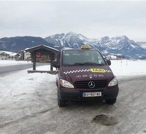 Ein Taxi steht auf einer schneebedeckten Straße mit einer malerischen Berglandschaft im Hintergrund. Die Umgebung ist winterlich und ruhig.