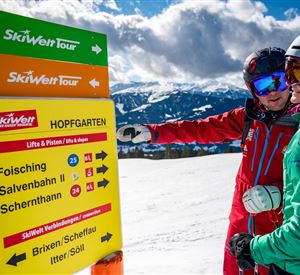 Een wintersporter wijst naar een bord met informatie over de SkiWelt Tour in Hopfgarten. Op de achtergrond zijn besneeuwde bergen en een blauwe lucht te zien.