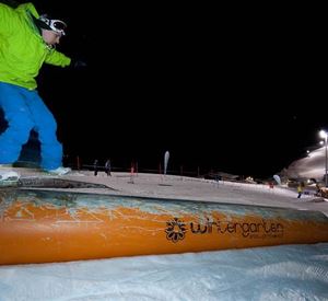 Ein Snowboarder springt über ein oranges Rail bei Nacht. Die Piste ist beleuchtet und bietet eine winterliche Atmosphäre.