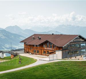Een grote houten berghut met een moderne aanbouw. Op de achtergrond zijn majestueuze bergen en een groene landschap te zien.