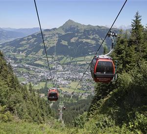 Eine Seilbahn fährt über eine grüne Landschaft mit Blick auf die Berge. Unten erstreckt sich ein malerisches Tal mit kleinen Häusern und viel Natur.