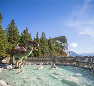 Ein großer Dinosaurier steht in einem glasklaren Wasserbecken, umgeben von Bäumen und Bergen. Der Himmel ist blau und sonnig.