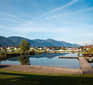 Ein ruhiger See umgeben von Bergen und Grün. Der Himmel ist klar und blau mit einigen Wolken.