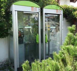 Two phone booths stand next to each other, surrounded by green plants. The phone booths have a modern shape and are partially equipped with glass fronts.