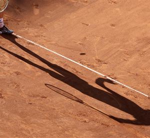 Ein Tennisspieler steht auf einem Sandplatz und spielt einen Ball. Der Schatten des Spielers ist deutlich auf dem Platz zu sehen.