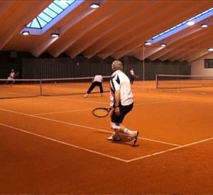 Een tenniswedstrijd in een hal met een oranje speelvloer. Meerdere spelers zijn actief aan het spelen en concentreren zich op het spel.