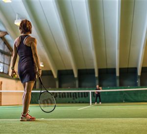 Een tennisster staat op het veld en kijkt naar het net. Het binnengebied is goed verlicht met een groene speelvloer.