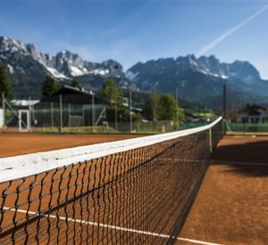 Een tennisbaan met rood zand en een heldere blauwe lucht. Op de achtergrond rijzen indrukwekkende bergen op.