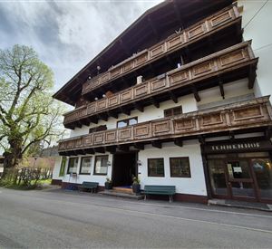 A traditional Alpine building with wooden balustrades and several floors. In front of the house is a quiet street and a large tree.