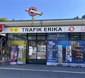 A small shop called "Trafik Erika" with a colorful range of products at the front. The surroundings are sunny, and there are advertising boards and magazines nearby.