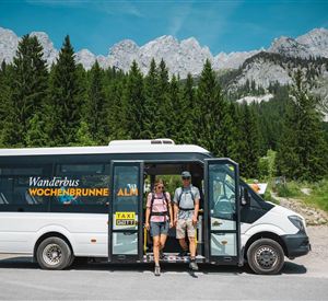 A hiking bus stands in nature, surrounded by mountains and forests. Two people get out, ready for an adventure.