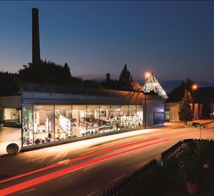 A modern building with large windows that is illuminated at night. In the foreground, there are streetlights and plants visible.