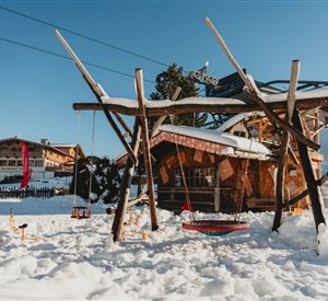 Zwei Personen in Winterkleidung stehen auf einer Eisbahn. Sie halten Besen und sitzen auf bunten Schlittenfiguren.