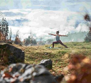 Een persoon doet yoga in een schilderachtige, bosrijke omgeving. Op de achtergrond zijn zachte heuvels en mist te zien.