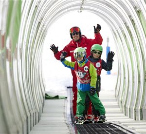 A ski instructor with two children in a ski tunnel. Everyone is smiling and waving at the camera.