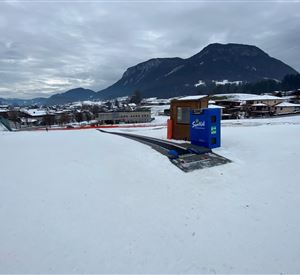 A snowy landscape with a small wooden house and a blue box. In the background, mountains and some houses can be seen.