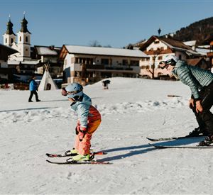 A family is skiing on a snowy slope. In the background, chalets and a church can be seen.