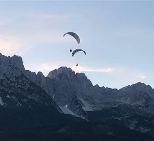 Two paragliders fly over majestic mountains. The sky is clear and showcases the beauty of nature.