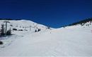 A snow-covered slope under a clear blue sky. In the background, ski lifts and trees are visible.