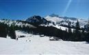 A beautiful winter landscape with snow-covered mountains and a clear blue sky. In the foreground, skiers on the slope and fir trees can be seen.