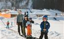 A group of children and adults is standing in a snowy area. They are wearing ski equipment and preparing to go down the slope.