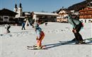 A child is learning to ski on a snow-covered slope. In the background, charming alpine huts and churches can be seen.