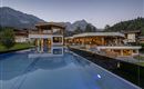 A modern building with large windows and a beautiful infinity pool. In the background, majestic mountains and trees are visible.