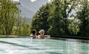 A couple relaxes in an infinity pool overlooking the mountains. The surroundings are green and the sun is shining.