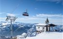 Eine schneebedeckte Berglandschaft mit einer Seilbahn und einem kleinen Gebäude. Der Himmel ist klar und blau, mit beeindruckenden Bergen im Hintergrund.