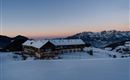 A rustic mountain cabin in the snow in front of majestic mountains. The sky displays soft colors at sunset.