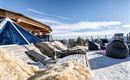 Eine gemütliche Terrasse mit Liegen und Sitzsäcken, umgeben von Schnee. Im Hintergrund sind klare Himmel und Berggipfel zu sehen.