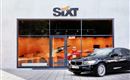 Ein modernes Mietwagenbüro von Sixt mit großen Fenstern. Vor dem Gebäude parkt ein schwarzes Auto.