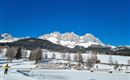 Eine winterliche Landschaft mit schneebedeckten Bergen und blauem Himmel. Im Vordergrund sind schneebedeckte Felder und Bäume zu sehen.