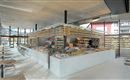 Ein modernes Café mit hellen, offenen Räumen und einer großen Theke, die verschiedene Lebensmittel anbietet. Die Einrichtung besteht aus Holz und bietet eine einladende Atmosphäre.