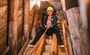 Eine Person sitzt in einem Minenschacht auf einer Holzrutsche und trägt einen gelben Helm. Der Raum ist aus Holz und beleuchtet, was eine spannende Atmosphäre schafft.
