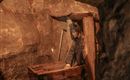 Ein Bergmann arbeitet in einem engen Tunnel. Im Hintergrund sind Werkzeuge und eine Holzkiste zu sehen.