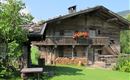 Ein traditionelles Holzhaus mit einem Balkon und roten Blumen. Der Garten ist grün und gepflegt, umgeben von Bergen.