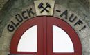 Das Bild zeigt eine rote Tür mit der Aufschrift „GLÜCK -AUF!“ und zwei gekreuzten Hämmern darüber. Die Tür ist Teil eines historischen Gebäudes und vermittelt ein handwerkliches Ambiente.