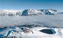 Eine schneebedeckte Berglandschaft mit majestätischen Bergen im Hintergrund. In der Mitte befindet sich eine Hütte, umgeben von Wolken und Skigebieten.