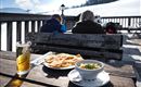 Ein Tisch im Freien mit Blick auf verschneite Berge. Darauf steht ein Glas Bier, ein Teller mit Fladenbrot und eine Schüssel mit warmem Gericht.