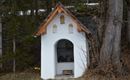 Eine kleine, weiße Kapelle mit einem Holzdach steht im Wald. Umgeben von Bäumen ist sie an einer ruhigen Stelle platziert.