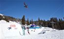 Ein Snowboarder springt in die Luft über einen verschneiten Park. Im Hintergrund sind Bäume und ein Skilift sichtbar.