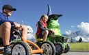 Two children are riding small vehicles on a sunny meadow. In the background, a large green figure with a wizard hat can be seen.