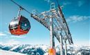 A cable car glides over a snowy mountain landscape. In the background, impressive mountains and a clear blue sky can be seen.