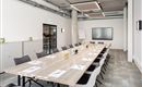 A modern meeting room with a long table and chairs.  
On the wall hangs a large screen, and there are various drinks and writing materials on the table.