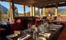 An elegant restaurant with wooden decor and red chairs. The table is festively laid with glasses and dishes, and large windows offer a view of the landscape.