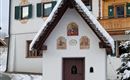 Eine kleine Kapelle mit einem schneebedeckten Dach und kunstvollen Fresken. Im Hintergrund sind traditionelle Holzhäuser zu sehen.