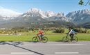 Two cyclists ride along a road on a vast green meadow. In the background, impressive mountains and a clear blue sky can be seen.