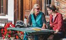 Two women are sitting at a table outdoors and are chatting. On the table, there is a bicycle helmet, a water bottle, and a banana.