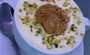 A cup with creamy drink, garnished with chopped pistachios. Next to the cup, there is a spoon on a saucer.